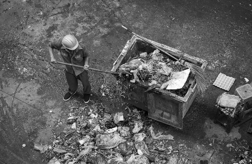 A person wearing a safety helmet and dark clothing is using a pitchfork to clear rubbish from the ground into a large metal skip situated on an outdoor paved area. The skip is filled with various waste materials, including debris and packaging, and is positioned beside a narrow wall or curb. Nearby, there are small scattered pieces of rubbish on the ground and a few objects such as a small stack of bricks or tiles. The environment appears to be an urban or industrial setting, possibly in Manor Park, with natural lighting highlighting the metallic finish of the skip and the textured surface of the waste materials. The scene reflects a professional rubbish removal process overseen by Office Clearance Manor Park, emphasizing the environmental cleanup and waste management service typical of local rubbish clearance operations.