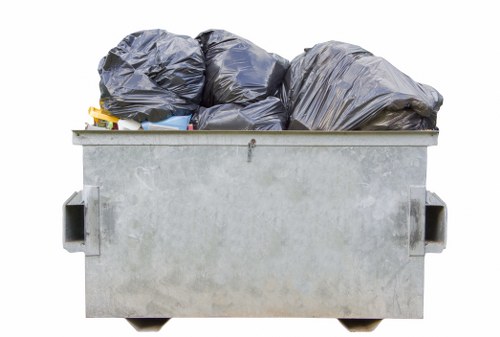 A large, rectangular metal skip with a light grey, slightly textured surface, situated outdoors on a paved area, filled with multiple tightly packed black garbage bags. The skip's side handles are visible on both sides, and the bags are piled unevenly, some leaning over the edges. The environment appears to be an open space possibly adjacent to a building or street, with no other objects or background details visible. The image conveys a scene of waste ready for collection, consistent with rubbish removal services offered by Office Clearance Manor Park near the local postcode area. The natural daylight illuminates the scene evenly, highlighting the industrial finish of the skip and the dark, plastic textures of the refuse bags, emphasizing the practical aspect of waste disposal activities in the local area.