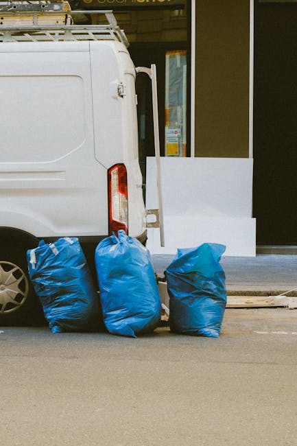 The image shows the rear end of a white commercial van parked on a paved street, with three large blue rubbish bags placed against the back wheel on the left side. The bags are made of plastic and app