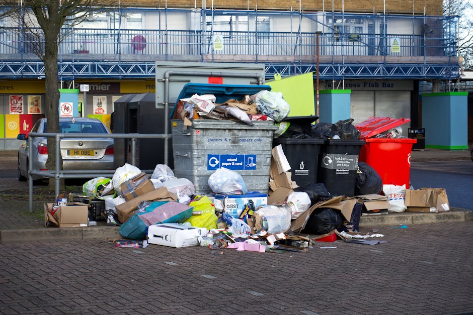 A collection of overflowing rubbish bins and scattered waste on a paved sidewalk in front of a commercial building with storefronts and a blue scaffolding structure. The waste includes black and red p