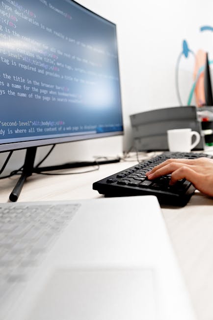 A person working at a desk in an office environment, viewed from behind with short dark hair and wearing a dark jacket. The individual is looking at a large computer monitor displaying lines of colorf